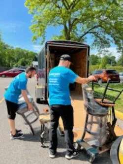 Two movers in blue shirts carrying chairs to a truck on a sunny street