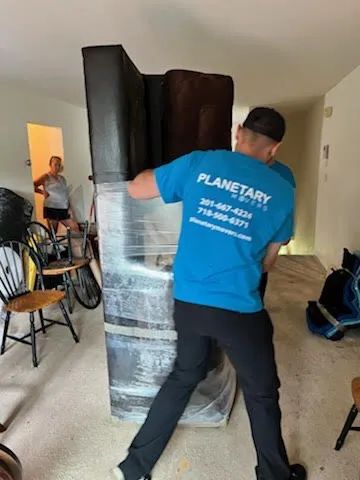 A Planetary Movers mover carrying furniture wrapped in plastic through an apartment